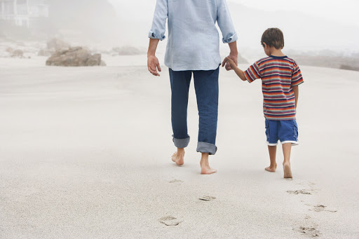 Father and son holding hands while walking on sand at beach &#8211; es