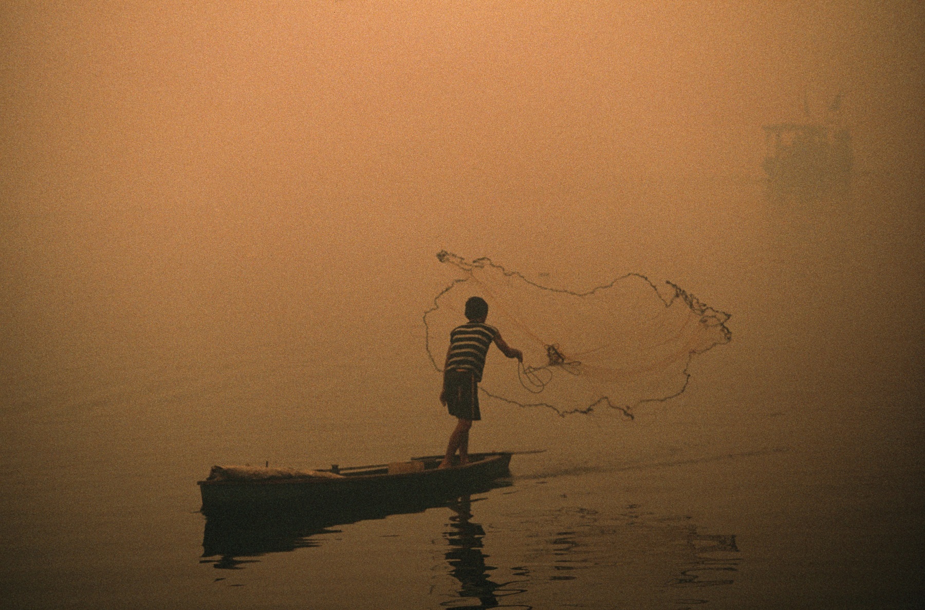 A boy casts his fishing net amidst smog from forest fires.