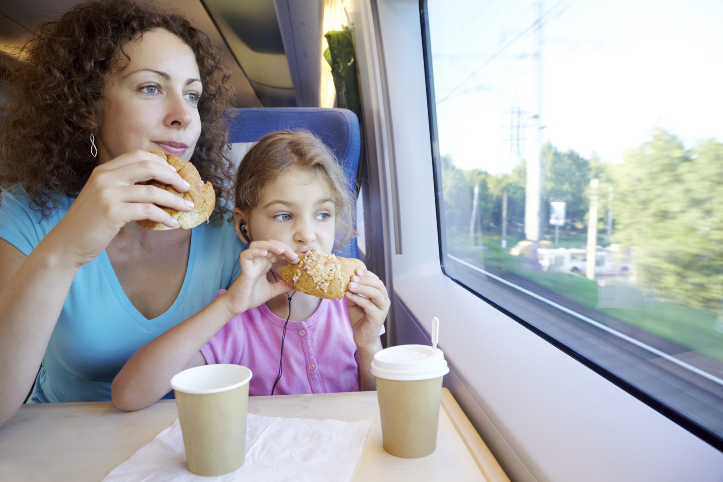 Mother and daughter eat and drink, sitting in armchair at table near window of moving speed train