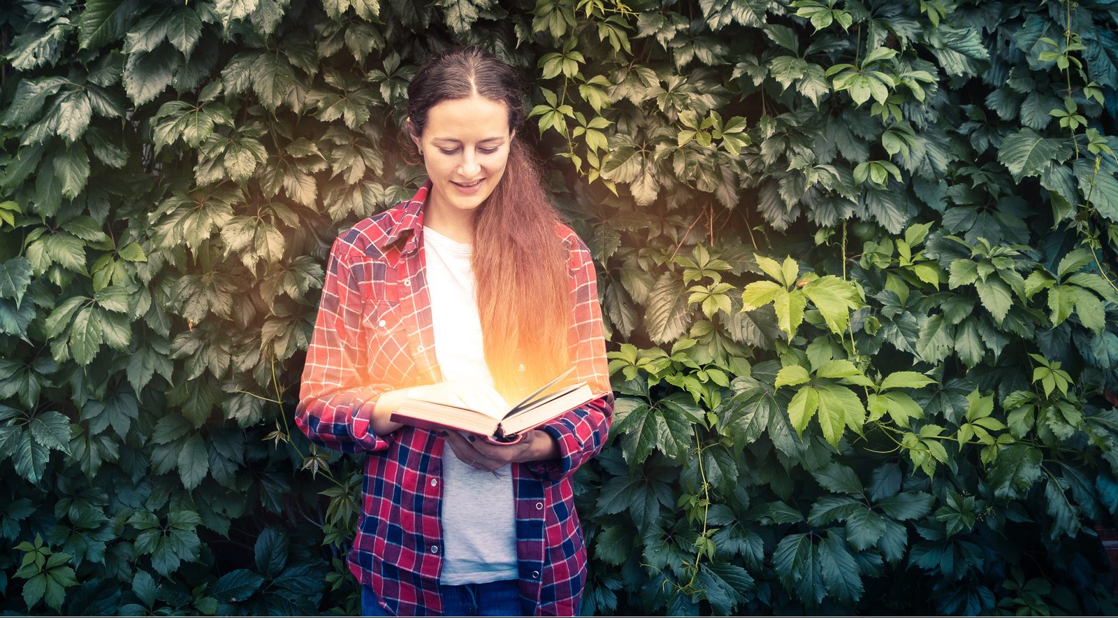 web2-young-dark-haired-woman-in-plaid-red-shirt-and-jeans-holds-in-her-hands-and-reads-a-magic-book-glowing-with-yellow-lights-against-the-beautiful-green-living-wall-of-the-grapes-shutt
