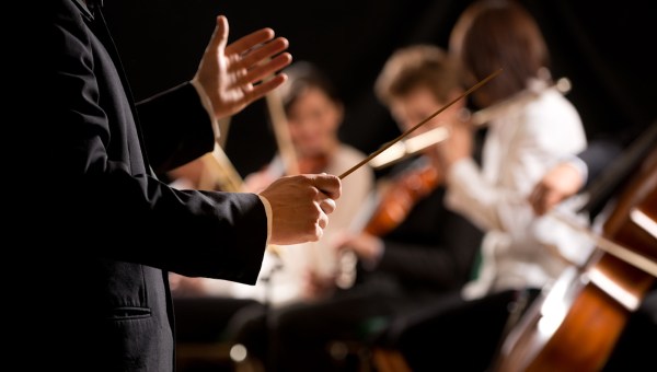 Maestro dirigindo orquestra sinfônica com artistas no fundo, mãos close-up.