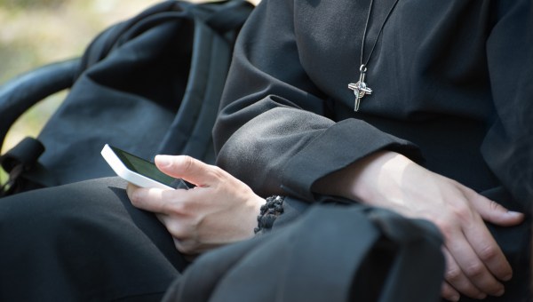 Unrecognizable-nun-in-a-black-robe-with-a-cross-on-his-neck-uses-a-smartphone-