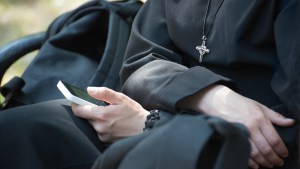 Unrecognizable-nun-in-a-black-robe-with-a-cross-on-his-neck-uses-a-smartphone-