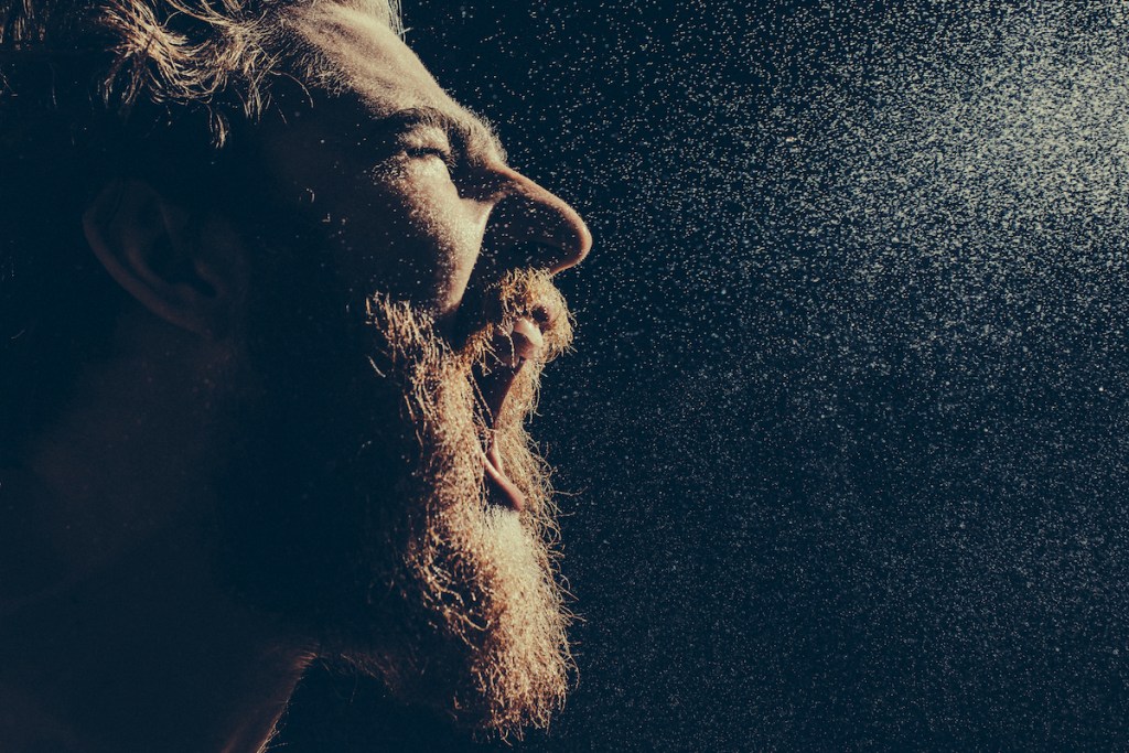 Naked bearded man angrily screams into a spray of water against a black background with copy space