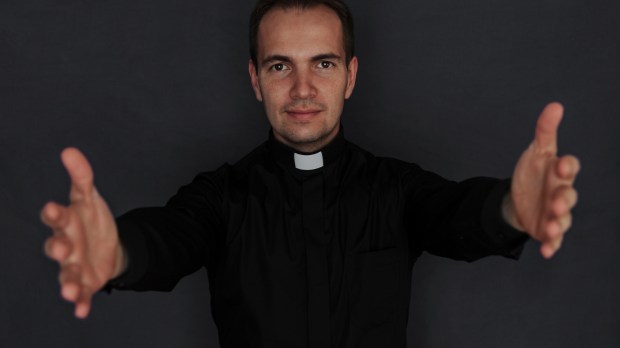 Młody ksiądz katolicki wyciąga ręce przed siebie stojąc na ciemnym tle