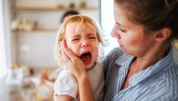 A mother holding a crying toddler daughter indoors in kitchen