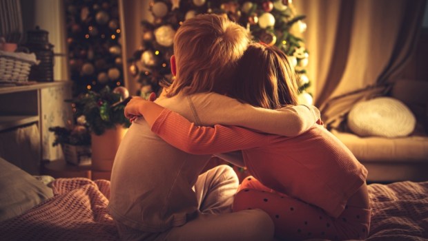 Children sit hugging in bed and look at the Christmas tree
